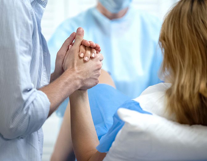 Man and woman holding hands in a hospital room, with a person wearing a mask in the background